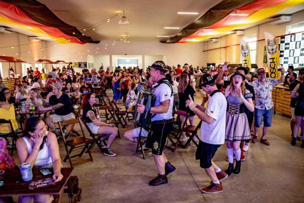 A imagem mostra um salão decorado com as cores da Alemanha durante uma festa temática. O local está cheio de pessoas sentadas em mesas e participando da celebração, enquanto músicos com trajes típicos se apresentam entre o público.