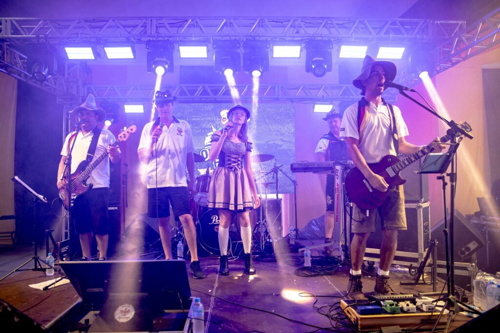 Os elementos da Banda Nostalgia atuam num palco ilumina por luzes roxas e holofotes, vestidos com trajes típicos alemães e chapéus característicos
