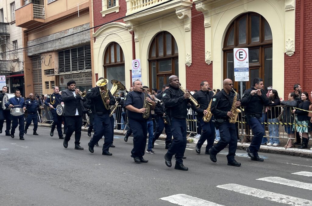 vemos uma banda marcial desfilando por uma rua da cidade, composta por músicos em uniformes escuros que tocam instrumentos de sopro como saxofones, trompetes, tubas e flautas, além de percussionistas com caixas. O desfile acontece diante de um prédio de tijolos vermelhos com detalhes claros e grandes janelas arqueadas, enquanto espectadores acompanham atrás de grades metálicas.