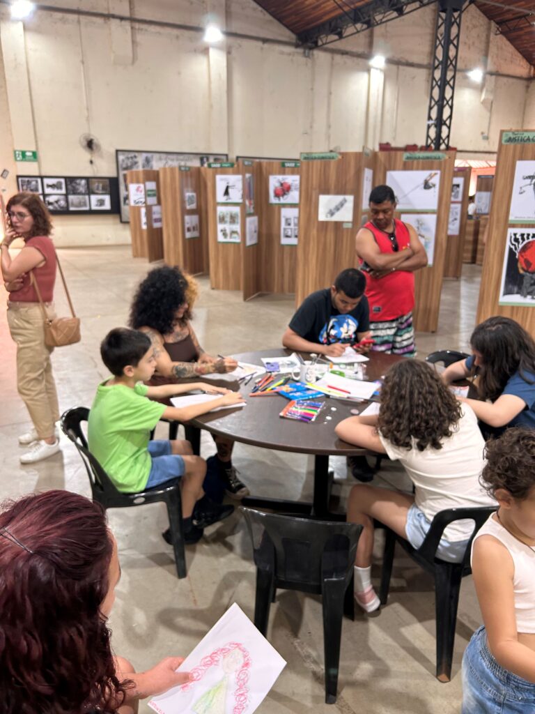 vemos um grupo de adultos e crianças reunidos em torno de uma mesa circular, envolvidos em atividades de desenho e pintura. O espaço está repleto de materiais artísticos como lápis de cor, canetinhas e folhas de papel, e uma das pessoas segura um desenho de uma figura com cabelo vermelho e vestido verde. Ao fundo, há painéis de madeira exibindo ilustrações e cartazes