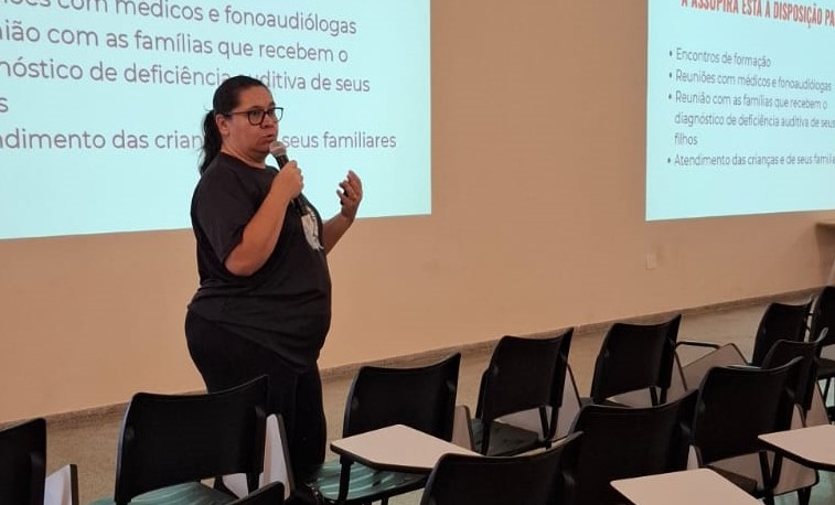 Uma mulher ministra uma palestra em uma sala de aula, falando ao público enquanto segura um microfone. Ao fundo, há projeções com textos e várias cadeiras escolares.