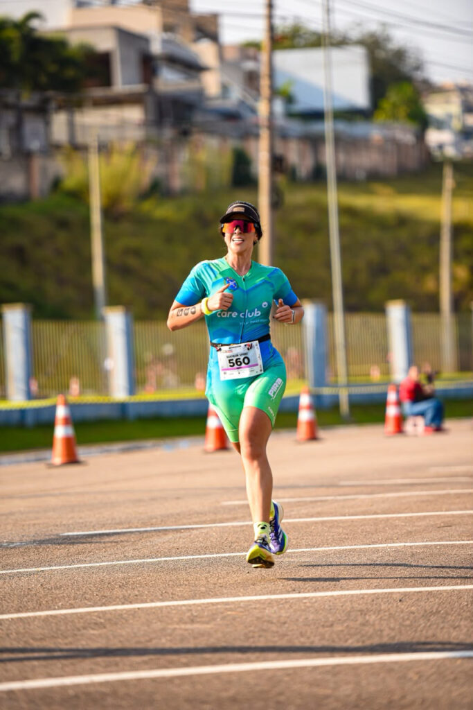 A imagem mostra uma pessoa participando de uma corrida em via asfaltada, usando uniforme esportivo azul e verde, tênis, boné e óculos esportivos.