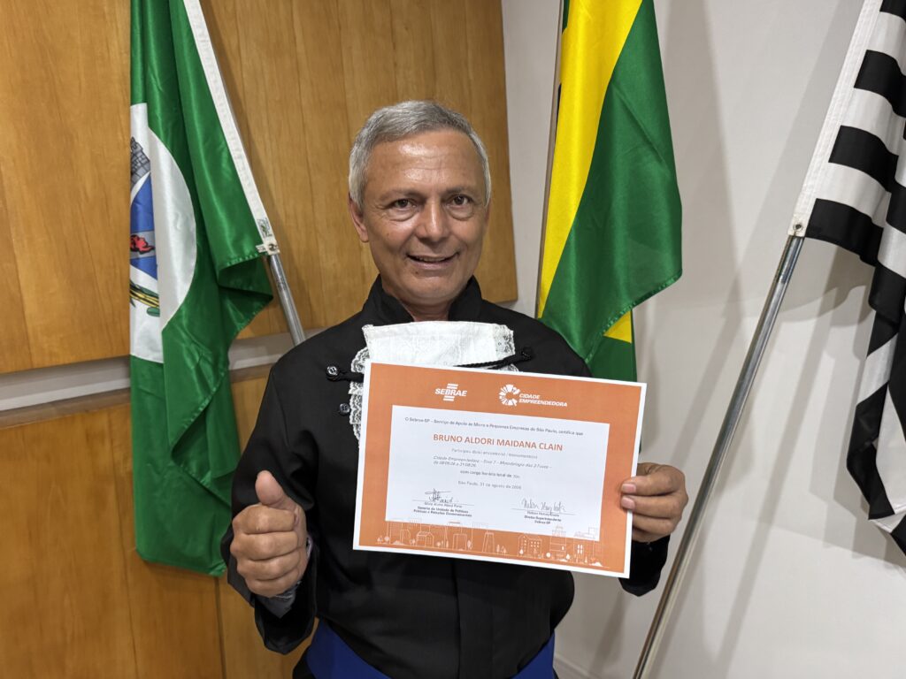 A foto mostra um homem sorrindo, beca preta. Ele segura um certificado emoldurado, com detalhes em laranja e os logotipos do Sebrae e do programa Cidade Empreendedora. No documento, aparece o nome Bruno Aldori Maidana Clain e a data 31 de agosto de 2026, em São Paulo. Ao fundo, há uma parede de madeira, uma bandeira do Brasil e parte de outra bandeira.