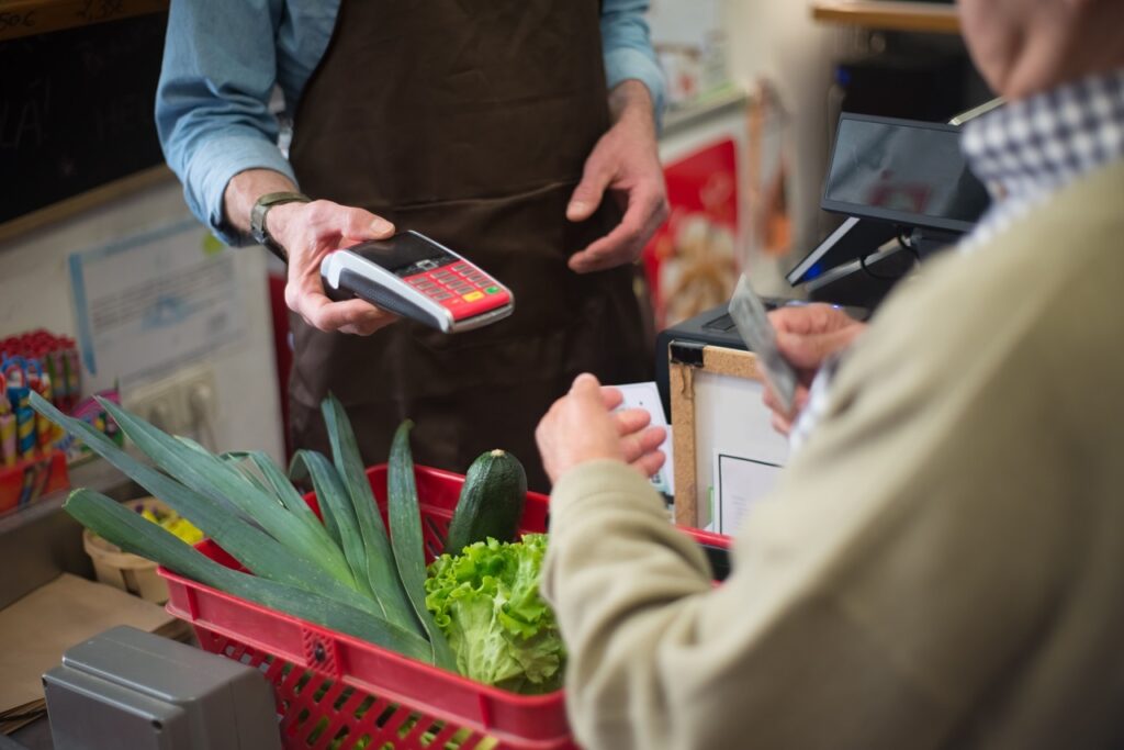 A imagem mostra uma pessoa no caixa de um mercado pagando suas compras. O atendente, usando avental escuro, segura uma máquina de cartão com teclado vermelho, enquanto o cliente segura dinheiro ou um cartão. No carrinho vermelho há vários vegetais frescos, como alho-poró, abobrinha e alface. Ao fundo, aparecem o monitor do caixa, produtos nas prateleiras e parte de um quadro-negro.