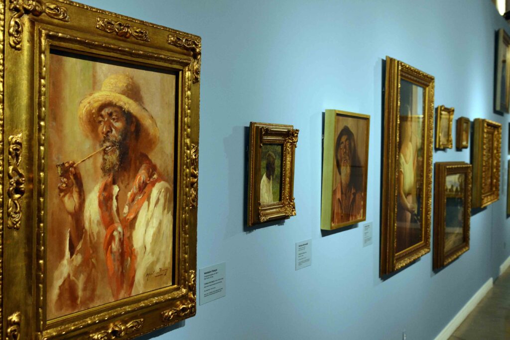 Galeria de arte com várias pinturas emolduradas em dourado expostas em uma parede azul, destacando um retrato de uma pessoa com chapéu.