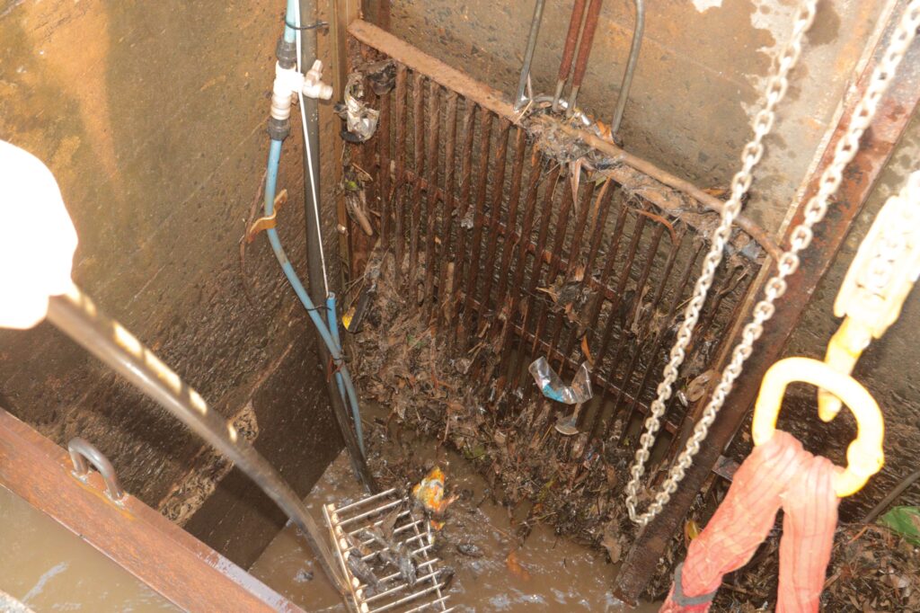 A imagem mostra o interior de uma câmara de drenagem ou poço de inspeção. As paredes são de concreto e há uma grade metálica parcialmente obstruída por detritos — folhas, plásticos e outros resíduos.