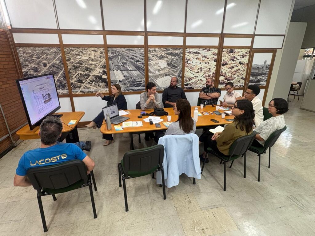 A imagem mostra um grupo de dez pessoas reunidas para uma foto em uma sala, diante de painéis com imagens históricas de uma cidade.