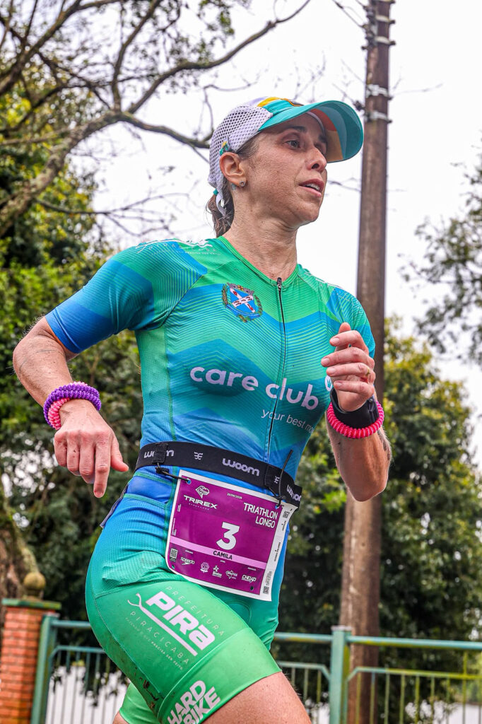 Atleta participa de uma prova de corrida, usando uniforme esportivo verde e número de identificação preso à roupa durante uma etapa de triatlo.