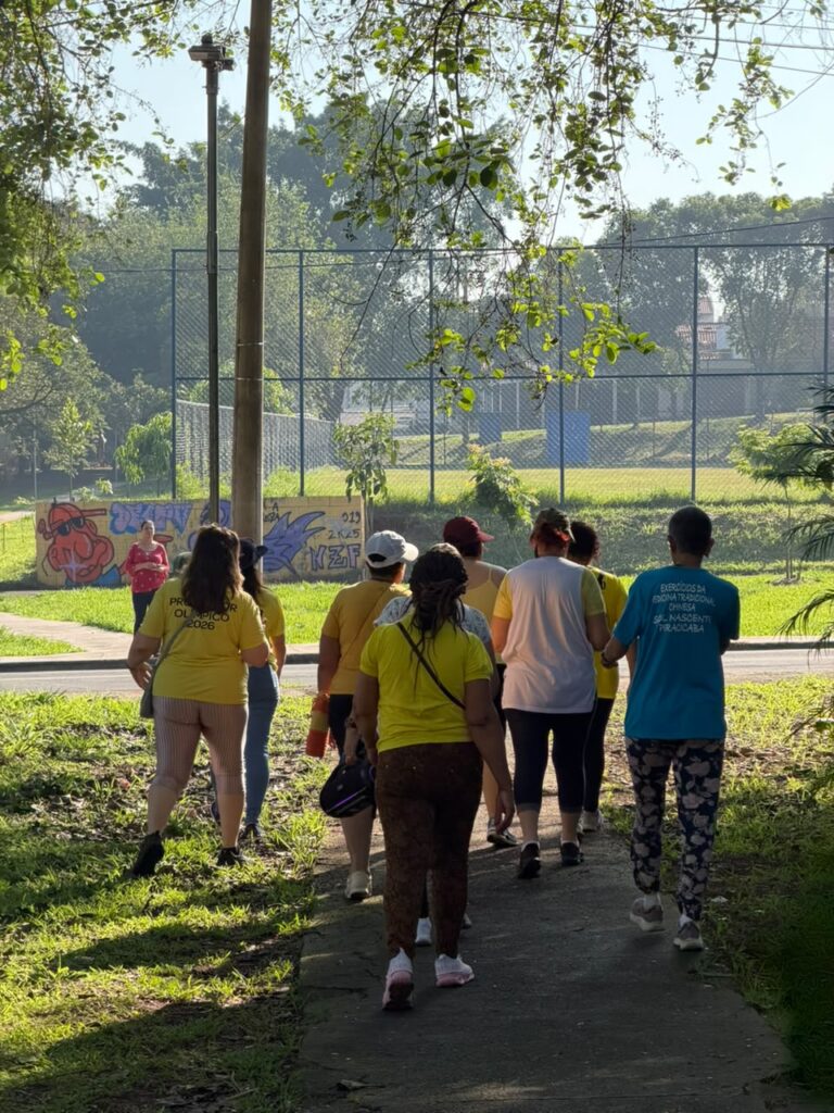 Grupo de pessoas participa de uma caminhada em uma área verde, seguindo por uma pista cercada por árvores e gramado, próxima a uma quadra esportiva.