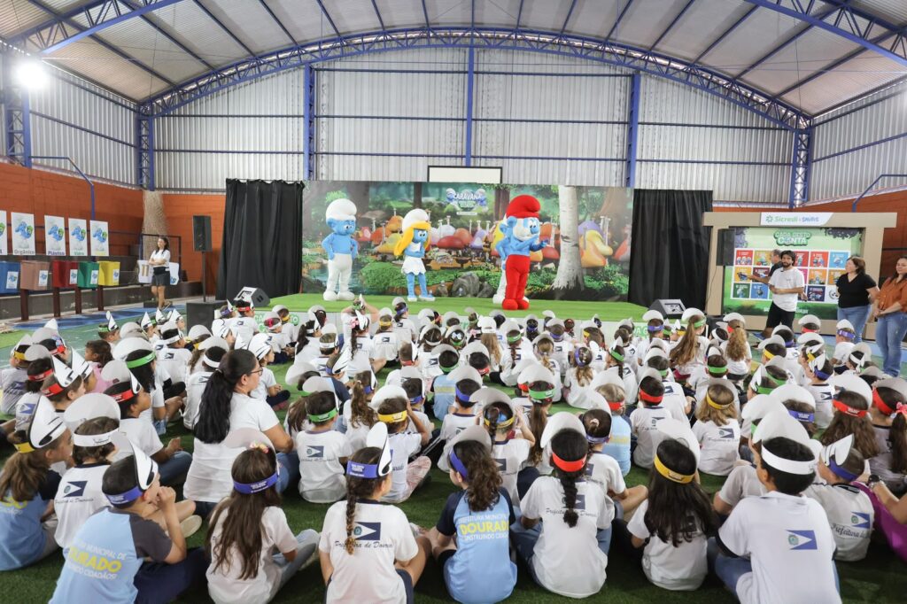 Um grande grupo de crianças da rede municipal, vestidas de t-shirt branca e com adereços na cabeça, encontra-se sentado no chão de um ginásio coberto a assistir a uma apresentação. No palco decorado ao fundo, estão três mascotes em tamanho real dos Smurfs (incluindo o Papai Smurf e a Smurfina) acompanhados por monitores do evento.