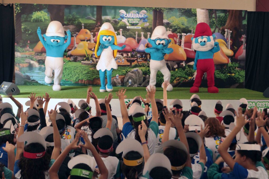 Personagem dos Smurfs em tamanho gigante interage com o público em um espaço repleto de estudantes participantes do evento.