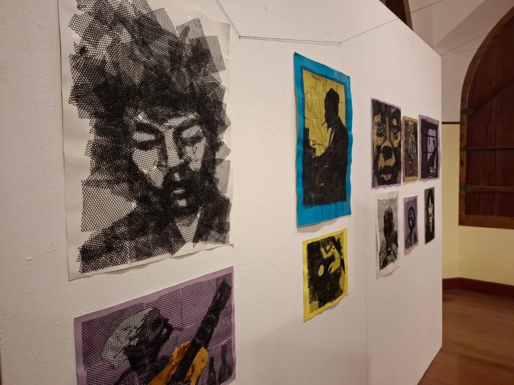 Galeria de arte com obras de estilo gráfico e retratos artísticos expostos em painéis brancos, incluindo imagens em preto e branco e composições coloridas.