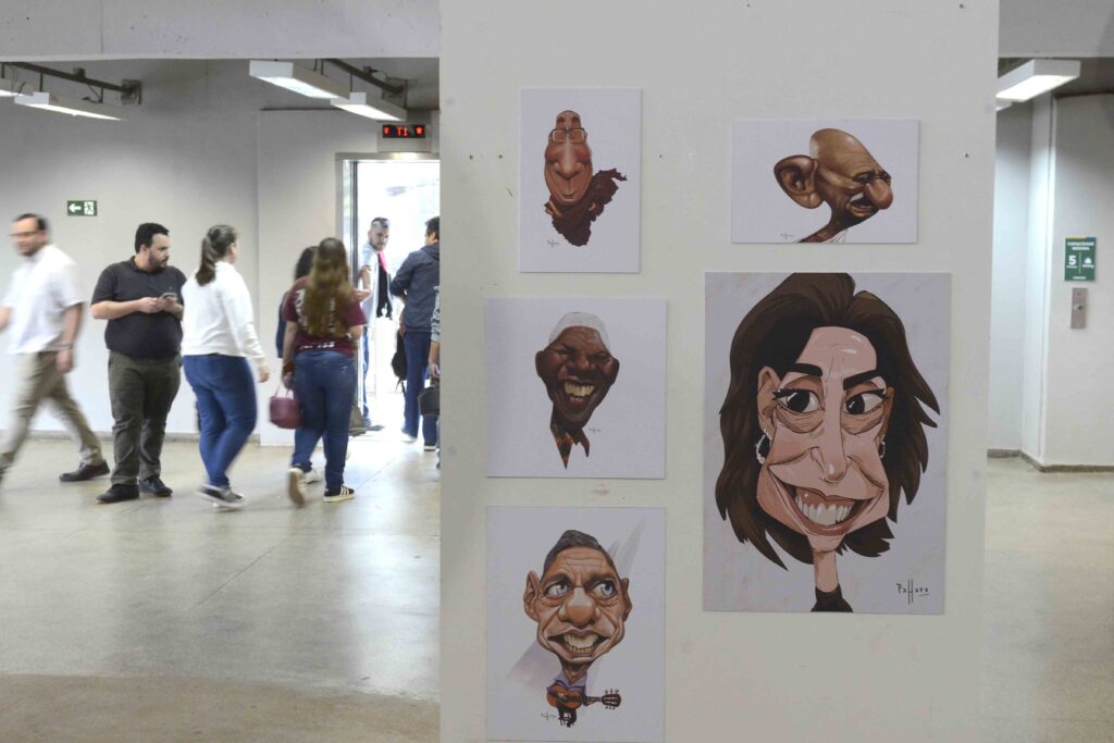 Painel de exposição com várias caricaturas e ilustrações artísticas fixadas em uma parede, enquanto pessoas circulam pelo espaço ao fundo.