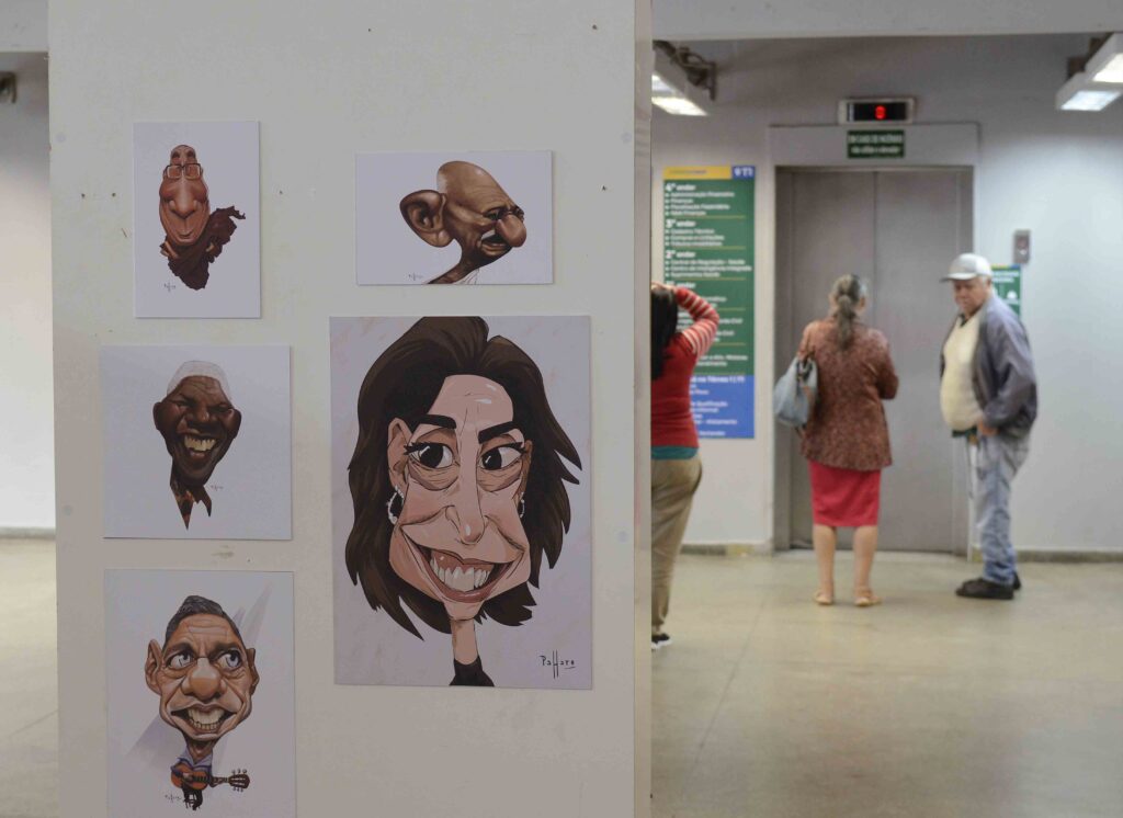 Mostra de caricaturas em um espaço cultural, com desenhos de personagens de traços exagerados expostos em um painel, enquanto visitantes circulam ao fundo.
