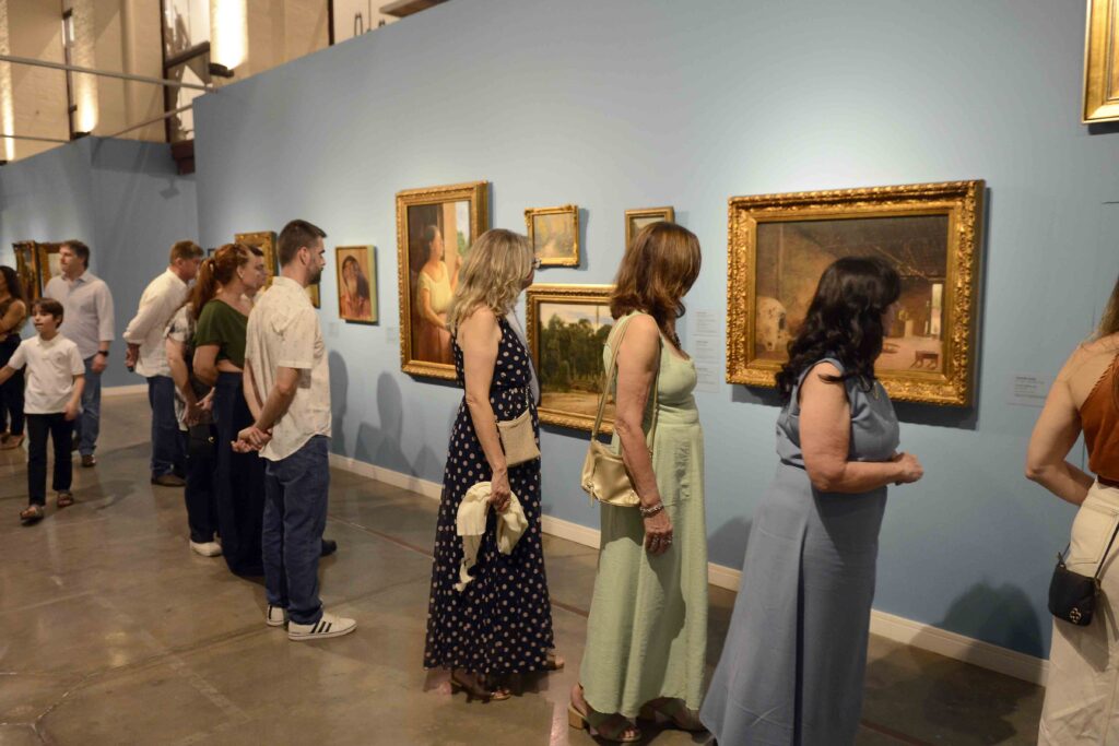 Visitantes observam pinturas emolduradas em uma galeria de arte com paredes azuis durante uma exposição.