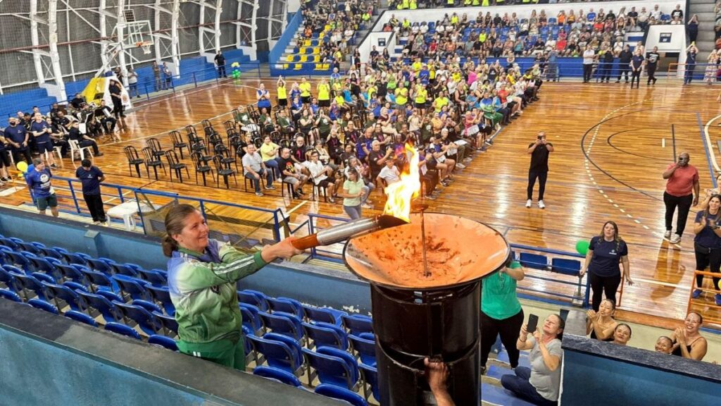 Uma mulher de agasalho verde acende a pira olímpica com uma tocha durante a cerimónia de abertura da Olimpíada da Melhor Idade, num ginásio repleto de público nas bancadas e na quadra.