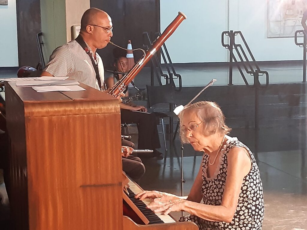 A imagem mostra uma apresentação musical em ambiente interno. Em primeiro plano, uma pessoa toca piano enquanto outra executa um fagote ao lado. O espaço possui iluminação suave e estrutura semelhante a um auditório ou sala de concertos, com outros músicos e estantes de partitura visíveis ao fundo.