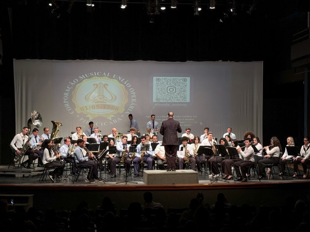 A imagem mostra uma banda musical se apresentando em um palco de teatro. Cerca de trinta músicos, vestidos principalmente com camisas brancas e calças escuras, estão sentados atrás de estantes com partituras e tocam instrumentos de sopro, como saxofones, trompetes, trombones, tubas e clarinetes. No centro, um maestro de costas para o público conduz a apresentação sobre um pequeno pódio. Ao fundo, há uma projeção com o nome “Corporação Musical União Operária – Piraçicaba”, um logotipo com uma lira e um código QR associado ao Instagram “@banda_uniaoop”. A plateia aparece em silhueta na parte inferior da foto.