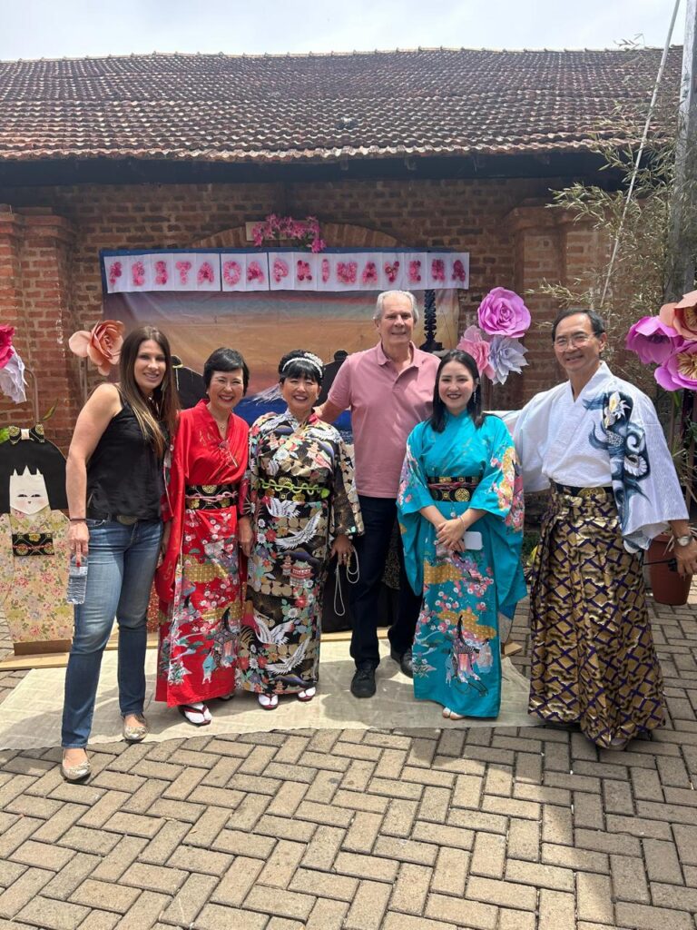 grupo de participantes posa em frente a uma decoração temática com flores e elementos da cultura japonesa. Algumas pessoas vestem quimonos coloridos, enquanto outras usam roupas casuais.