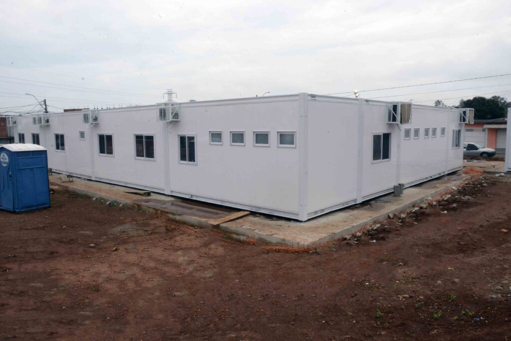 A imagem mostra uma construção modular com um banheiro portátil ao lado. A imagem exibe um conjunto de construções modulares brancas conectadas em um longo comprimento, prontas para uso ou em fase de instalação. As unidades possuem múltiplas janelas de diferentes tamanhos e instalações de ar condicionado montadas nas paredes externas. À esquerda da estrutura principal está um banheiro portátil azul, rotulado com "Ativa" e um logotipo associado. O terreno ao redor é um solo marrom, irregular, com algumas áreas de cimento e materiais de construção espalhados, como tábuas de madeira e uma rede laranja. O céu está nublado e cinza, criando uma atmosfera um pouco sombria e indicando um clima potencialmente chuvoso ou tempestuoso. A cena sugere um local de construção ou uma instalação temporária em andamento, talvez para eventos, acampamentos ou como alojamento de emergência. A ausência de pessoas e o estado do terreno conferem à imagem um sentimento de transitoriedade e de trabalho em progresso.