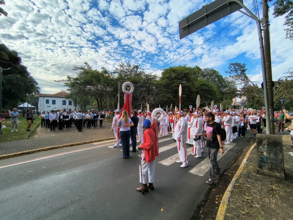 A imagem mostra uma procissão ou desfile religioso acontecendo em uma rua arborizada. Muitas pessoas vestem roupas brancas com detalhes vermelhos e carregam bastões decorados com grandes círculos brancos, além de estandartes vermelhos. À esquerda, há uma banda musical formada por vários integrantes com instrumentos de sopro. O público acompanha pelas calçadas, enquanto algumas pessoas fotografam ou filmam. Ao fundo, aparecem árvores, um prédio branco e uma paisagem urbana. O céu está azul, coberto por nuvens espalhadas.