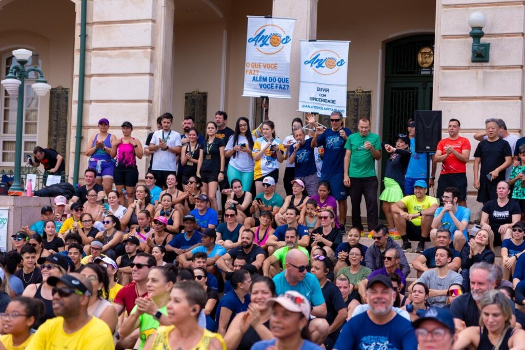 A imagem mostra uma grande multidão reunida em frente a um prédio histórico, provavelmente durante um evento esportivo ou ação comunitária. Muitas pessoas usam roupas coloridas, bonés e camisetas esportivas; algumas estão sentadas nos degraus, enquanto outras permanecem em pé e aplaudem. No alto, há dois banners com o logotipo “Anjos” e as frases: “O que você faz além do que você faz?” e “Ouvir com sinceridade”. Também aparecem caixas de som e pessoas segurando microfones, indicando que há uma apresentação ou cerimônia acontecendo.