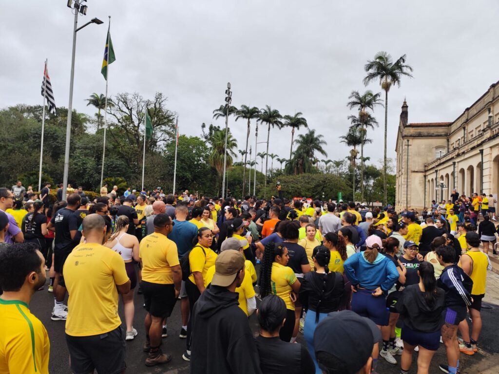 Uma grande multidão de participantes de uma corrida de rua se reúne em uma praça pública ao ar livre, aguardando o início do evento em uma manhã nublada de domingo. A composição da cena é aberta e dinâmica, apresentando uma aglomeração densa de pessoas distribuídas por toda a área central da imagem. O enquadramento em plano geral captura a interação casual dos corredores, que conversam entre si, alongam-se ou caminham em direção à linha de partida. O posicionamento dos indivíduos é orgânico, criando um fluxo natural que se estende desde o primeiro plano até o fundo, onde a multidão se dispersa em direção às árvores e ao edifício histórico à direita. A disposição dos corredores sugere um momento de pré-prova, com uma atmosfera de expectativa coletiva que domina o espaço. Os sujeitos principais são homens e mulheres de diversas idades, majoritariamente vestidos com roupas esportivas, como camisetas de corrida de cores vibrantes — especialmente amarelas, pretas e azuis — além de shorts e tênis de performance. Algumas pessoas usam bonés e acessórios esportivos, com expressões faciais variando entre a descontração, o foco na preparação física e a socialização. A diversidade de trajes, incluindo algumas vestimentas casuais, reflete o caráter inclusivo do evento, com os participantes mantendo posturas variadas: alguns em pé, outros orientados para os amigos e alguns caminhando pelo local. Como se trata de uma fotografia documental, a técnica foca no registro realista do evento. A execução é direta, mantendo um foco nítido em toda a cena e utilizando uma profundidade de campo que permite visualizar tanto as pessoas próximas quanto a arquitetura e a vegetação ao fundo. Não há manipulações estilísticas, priorizando a autenticidade do momento, com o enquadramento equilibrando a massa de pessoas com elementos ambientais significativos, como as bandeiras hasteadas e a construção clássica que serve como um marco visual. O cenário é um espaço aberto que combina elementos urbanos e naturais, como um pátio pavimentado adjacente a uma estrutura arquitetônica clássica com arcadas e janelas tradicionais. Ao fundo, palmeiras altas e outras árvores compõem a paisagem, reforçando uma atmosfera tropical. As condições de iluminação são marcadas por um céu nublado, que oferece uma luz suave e difusa, eliminando sombras fortes e criando uma claridade uniforme sobre toda a multidão. O clima geral é de tranquilidade e energia comunitária, típico de eventos esportivos matinais onde o entusiasmo dos participantes contrasta com a calma do céu nublado.