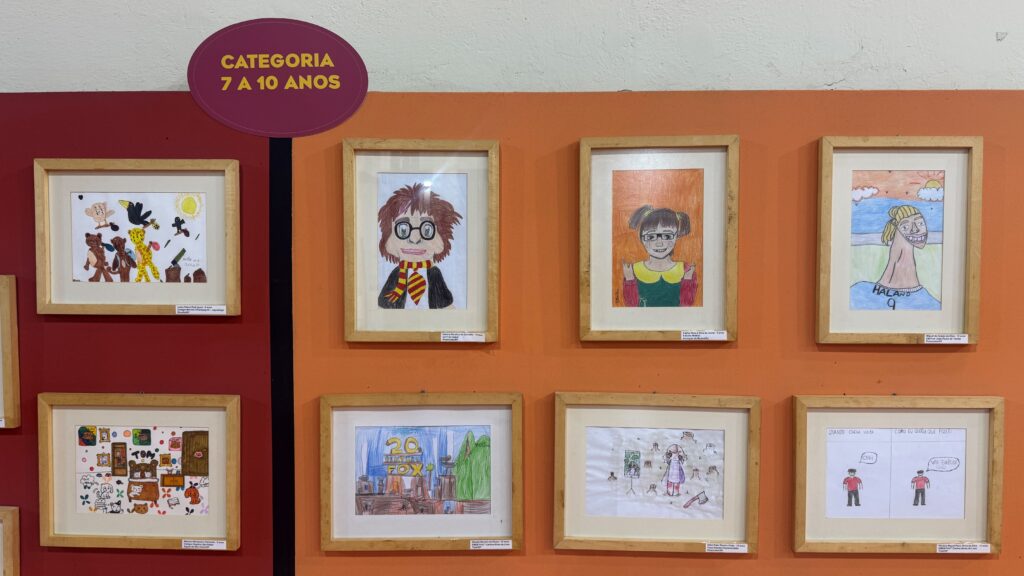 A imagem mostra uma parede de exposição com vários desenhos infantis emoldurados. No topo, há uma placa com o texto “Categoria 7 a 10 anos”, indicando a faixa etária dos participantes.
