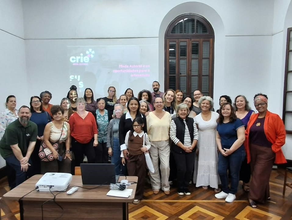 A imagem mostra um grupo grande de pessoas reunido em uma sala, posando para uma foto. Ao fundo, há uma projeção com o logotipo “crie” e o texto “Moda Autoral: oportunidades para o artesanato”. As pessoas estão diante de uma parede clara, com uma janela alta de madeira. Em primeiro plano, há uma mesa com um projetor, um notebook, um controle remoto e alguns materiais. O piso tem um padrão geométrico de madeira.