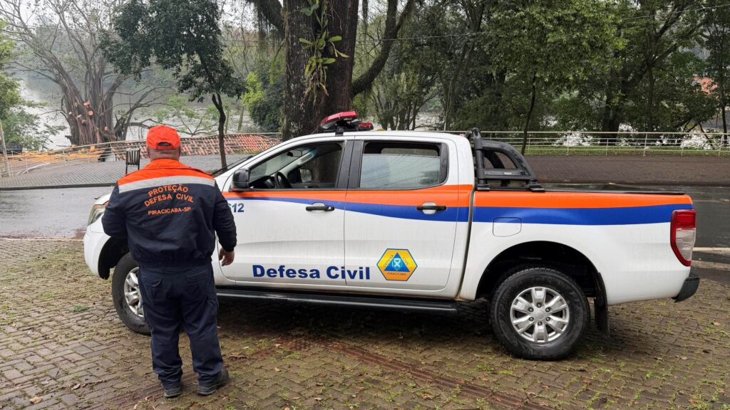 A imagem mostra uma viatura da Defesa Civil estacionada em uma área próxima a um rio ou lago, em um dia chuvoso. Ao lado do veículo, uma pessoa uniformizada observa o local.