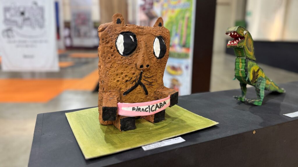 A imagem mostra uma exposição de esculturas artesanais. Em destaque, há uma figura marrom com grandes olhos pretos e brancos, textura rugosa e uma faixa rosa com a palavra “PIRACICABA”. Ao lado, encontra-se uma pequena escultura de dinossauro verde com a boca aberta e dentes aparentes.