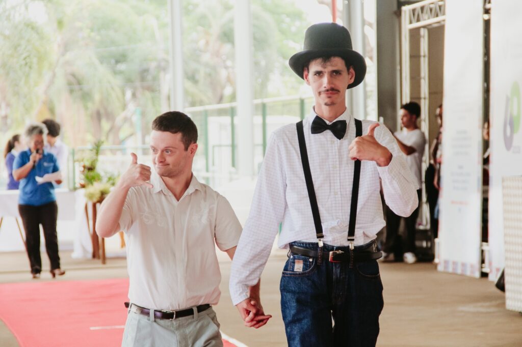 Copilot said: A imagem mostra duas pessoas caminhando de mãos dadas em uma passarela com tapete vermelho durante um evento. Uma delas usa camisa branca, suspensórios, gravata borboleta e cartola, enquanto a outra veste roupa clara.