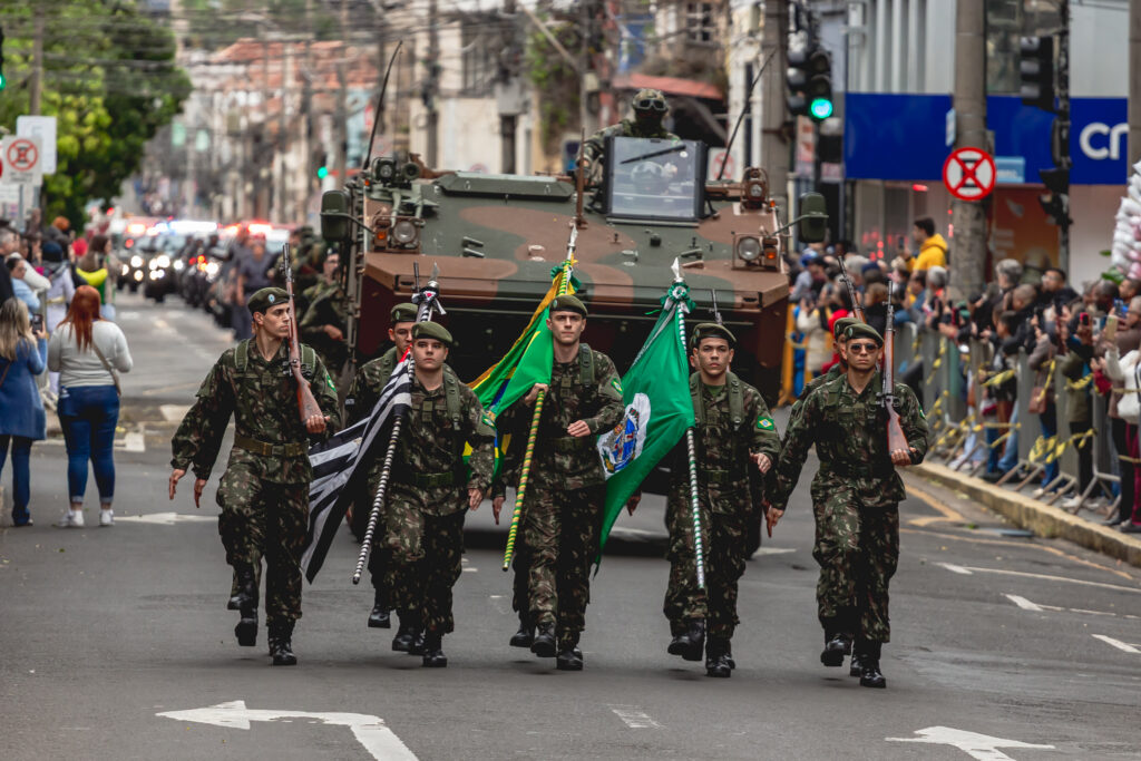 A imagem mostra seis soldados em uniforme camuflado e boinas verdes marchando em formação. Dois deles carregam rifles, enquanto outros exibem bandeiras — entre elas a do Brasil, a do estado de São Paulo e uma bandeira verde com emblema. Logo atrás, um veículo blindado acompanha a tropa, com um militar visível pelo escotilhão. O público assiste dos dois lados da via, separado por barreiras e fitas amarelas, compondo uma cena solene e organizada que transmite disciplina e patriotismo em um evento cívico.