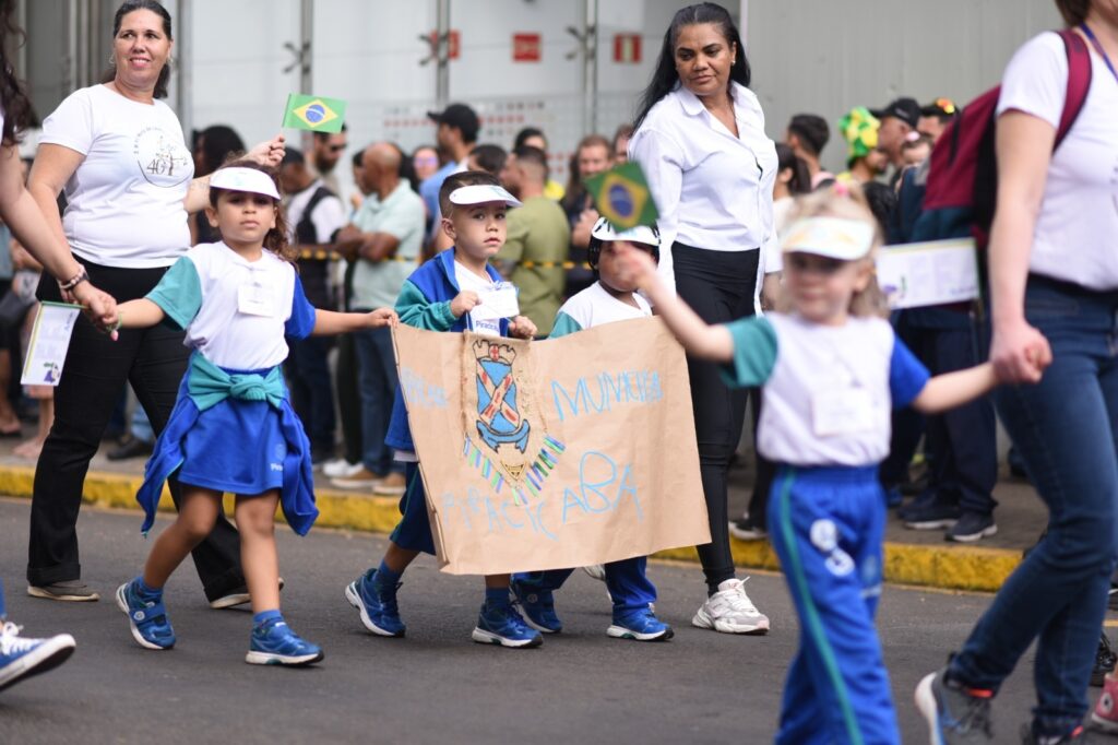 A imagem mostra um desfile ou apresentação infantil em uma rua. Várias crianças usam uniformes nas cores azul, verde e branco, com viseiras brancas, enquanto caminham segurando um cartaz de papelão. No cartaz aparece um brasão e o texto “Municipal Piracicaba”. Algumas crianças também carregam pequenas bandeiras do Brasil. Adultos acompanham o grupo, e ao fundo há uma multidão de espectadores diante de um grande prédio.