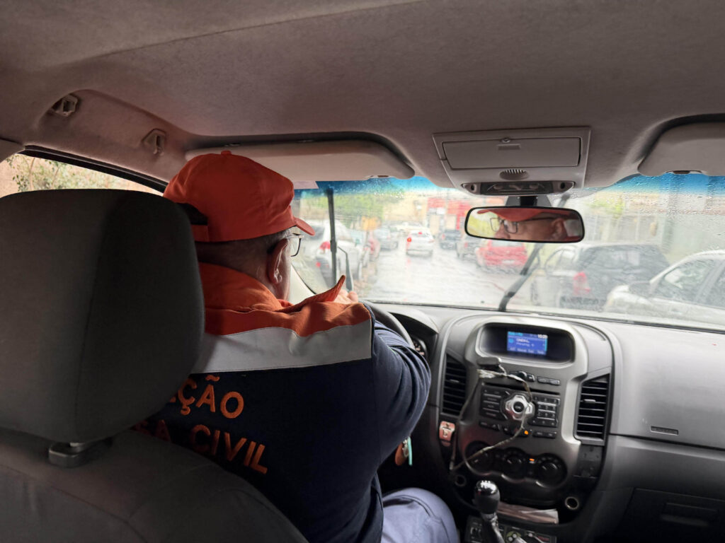 A imagem mostra o interior de um veículo visto do banco traseiro, com um agente da Defesa Civil ao volante. Ele usa boné e uniforme laranja e azul, e parece concentrado na direção enquanto percorre uma rua urbana em dia chuvoso — o para-brisa está molhado e os limpadores em movimento. O cenário sugere uma ação de monitoramento ou vistoria realizada pela equipe durante condições de chuva.