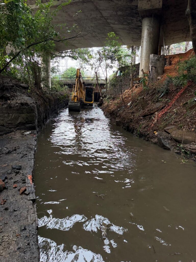 A imagem mostra um canal estreito com água barrenta passando sob uma ponte de concreto. No centro do canal, há uma escavadeira realizando trabalhos de limpeza ou desobstrução do leito.