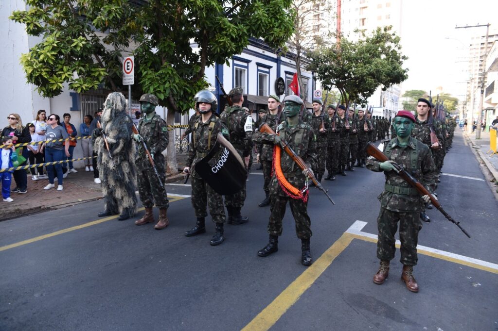 A imagem mostra um desfile militar em uma rua urbana. Vários soldados usam uniformes camuflados, boinas ou capacetes e carregam rifles; alguns têm o rosto pintado de verde. Um deles segura um escudo preto com a palavra “CHOQUE”, enquanto outro veste uma roupa de camuflagem especial, semelhante a um traje de folhagem. Ao lado da rua, espectadores observam atrás de uma fita de isolamento. Há árvores, prédios, placas de trânsito e barreiras amarelas, indicando que a via foi parcialmente fechada para o evento.