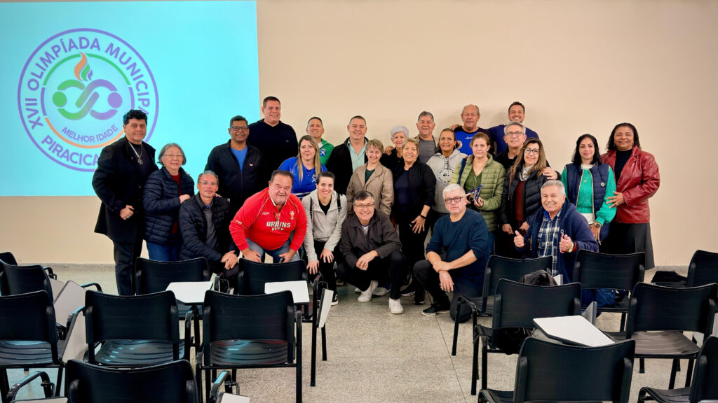 A imagem mostra um grupo de cerca de 25 pessoas reunidas em uma sala, posando para foto diante de uma projeção com o logotipo da XVIII Olimpíada Municipal da Melhor Idade – Piracicaba/SP.