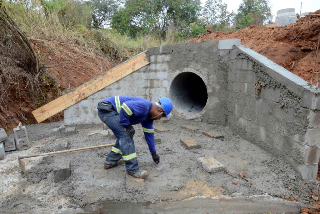 O trabalhador de capacete azul está moldando o concreto em torno de um grande tubo de drenagem. A estrutura faz parte de um sistema de escoamento de água em construção, com formas de madeira e blocos ao redor. O cenário mostra uma etapa essencial da engenharia civil voltada para infraestrutura e gestão hídrica.