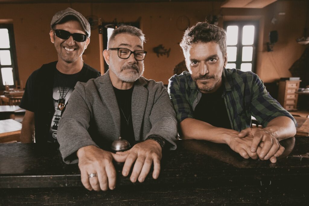 Três integrantes da banda Hotel Magnólia posam debruçados sobre um balcão de madeira num ambiente acolhedor, no âmbito da divulgação para a 3ª Oktoberfest de Piracicaba