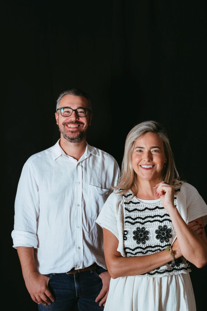 duas pessoas posam para uma foto em estúdio, diante de um fundo escuro, usando roupas claras e em composição voltada para divulgação artística.