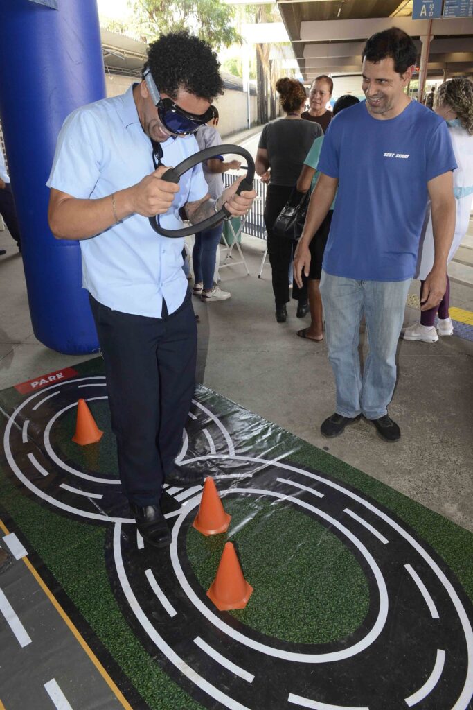 Um homem com óculos simuladores de embriaguez tenta caminhar sobre um tapete com marcações de pista de trânsito e cones de sinalização enquanto segura um volante, sob o olhar de outra pessoa.