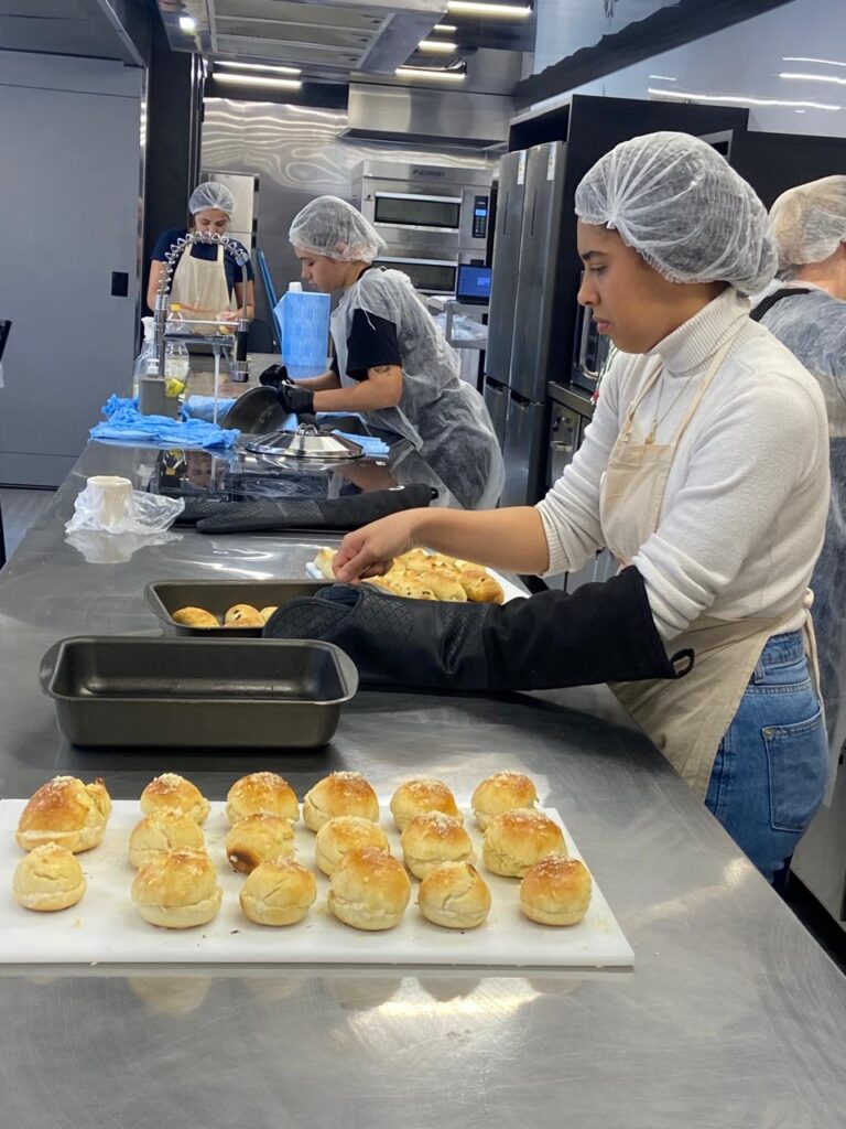 A imagem mostra uma aula de panificação em uma cozinha moderna e bem equipada. Quatro pessoas, todas usando toucas e aventais, trabalham em bancadas de aço inox, o que indica um ambiente profissional e higiênico.