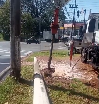 A imagem mostra uma equipe realizando uma obra de manutenção em uma área gramada, ao lado de uma avenida. No centro, um equipamento hidráulico vermelho perfura ou remove o solo, deixando uma abertura com terra e lama ao redor. À direita, parte de um caminhão de serviço aparece estacionada, com mangueiras e estruturas metálicas. Um tubo branco se estende pelo gramado em direção à área de trabalho. Ao fundo, há uma rua com faixa de pedestres, veículos, postes, fios elétricos e árvores.