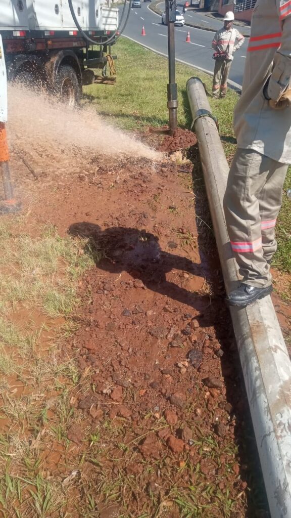 A imagem mostra trabalhadores realizando um serviço de manutenção em uma tubulação próxima a uma rodovia. Um caminhão está conectado a uma mangueira grossa, que lança água ou outro líquido sob pressão sobre o solo, formando uma grande poça de lama. Há um tubo metálico exposto ao lado da área escavada, enquanto um trabalhador permanece sobre ele e outro observa o serviço. Cones laranjas sinalizam a operação, e veículos passam pela estrada ao fundo.