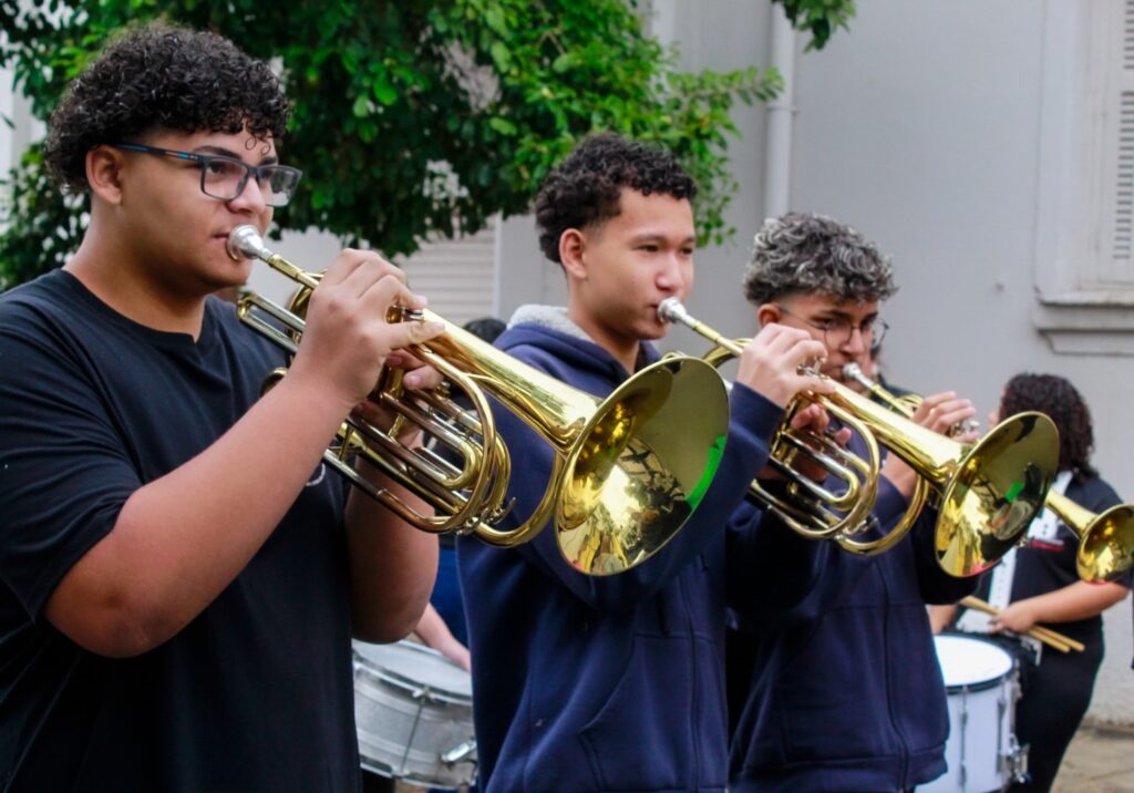 A imagem mostra um grupo de jovens músicos tocando instrumentos de metal, semelhantes a cornetas ou flugelhorns, durante uma apresentação ao ar livre. Em primeiro plano, três pessoas seguram os instrumentos dourados e os levam à boca para tocar.