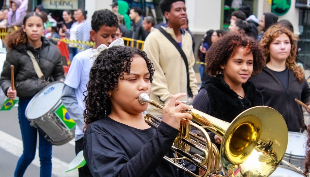 Copilot said: A imagem mostra uma apresentação de banda em uma rua movimentada. Em primeiro plano, uma criança toca um instrumento de sopro de metal dourado, enquanto outras crianças e jovens participam da apresentação com instrumentos de percussão ao fundo.