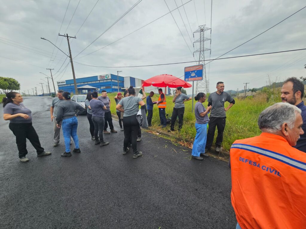 A imagem mostra um grupo de pessoas reunidas em uma rua asfaltada próxima a um prédio industrial azul e branco com o nome “Triton” e “Grupo Jacto” na fachada. A maioria veste camisetas cinza e calças escuras, enquanto um homem usa um colete laranja e outro veste uma jaqueta da Defesa Civil. Há também um guarda-sol vermelho montado à beira da estrada, perto de uma placa que indica “Ponto de Encontro (emergência)”. O clima parece nublado, sugerindo um dia de treinamento ou simulação de segurança.