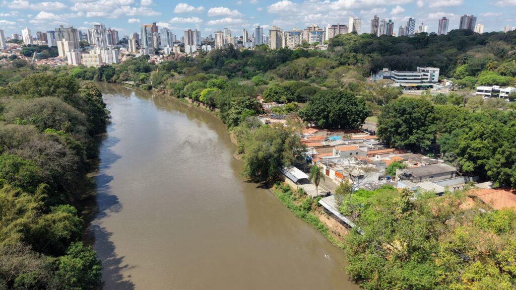 A imagem mostra uma vista panorâmica de uma cidade brasileira. Em primeiro plano, há um rio largo de águas barrentas, cercado por muita vegetação e árvores. À direita, aparecem casas simples, ruas e alguns edifícios comerciais próximos às margens. Ao fundo, destaca-se uma área urbana com muitos prédios altos de diferentes estilos e alturas. O céu está azul, com nuvens brancas, e a paisagem combina uma grande área verde com a intensa ocupação urbana.