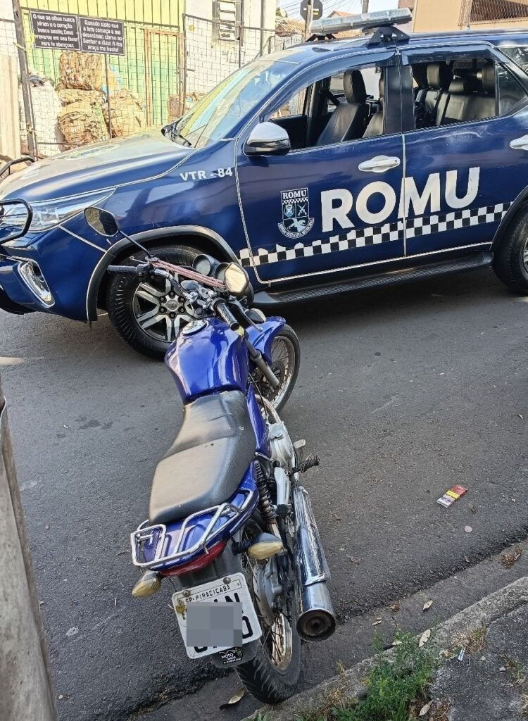 A imagem mostra uma motocicleta azul estacionada em primeiro plano, vista principalmente pela traseira. Ela tem banco preto, escapamento cromado, bagageiro metálico e placa parcialmente ocultada. Ao fundo, há um carro azul-escuro da ROMU, identificado como “VTR-84”, com faixas quadriculadas nas laterais e uma barra de luz no teto. Os veículos estão em uma rua asfaltada, diante de um portão e de uma parede com cartazes religiosos.